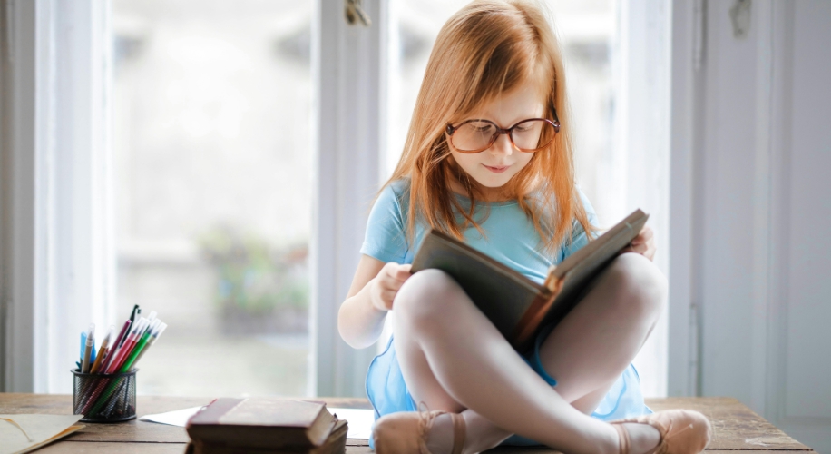 Child wearing glasses reading a book with legs crossed on a table.