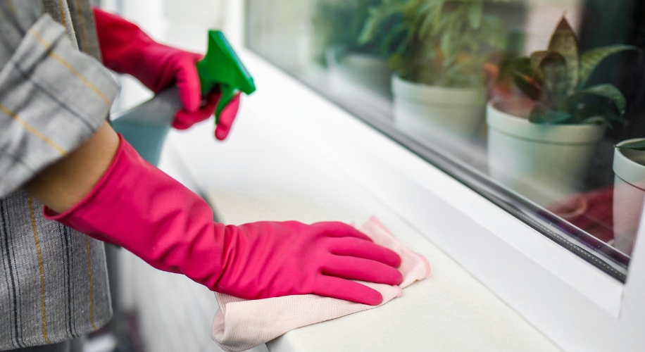 Hands wearing pink rubber gloves cleaning a windowsill and spraying cleaner with a green spray bottle.