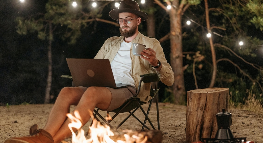 Man using a laptop while holding a mug, sitting on a camping chair by a campfire at night.