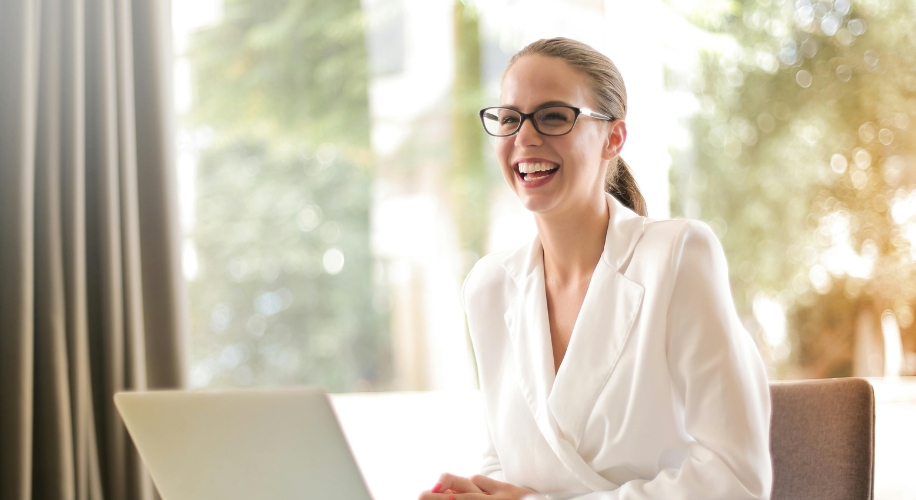 Woman in a white suit, smiling and wearing glasses, sitting in front of a laptop.