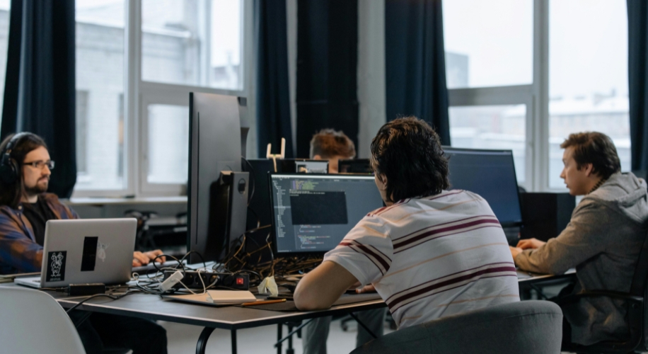 People working at computers in an office setting. Multiple monitors show coding and development tools.