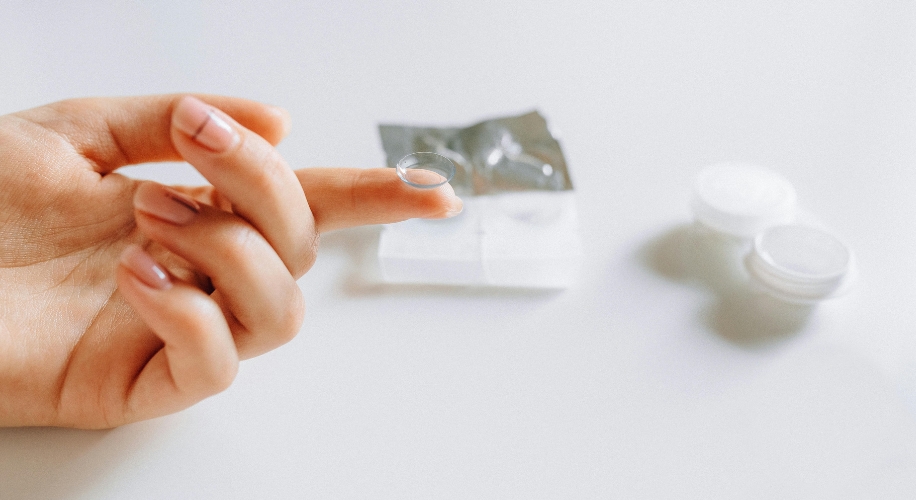 Contact lens balanced on fingertip, with open contact lens case and foil package in the background.