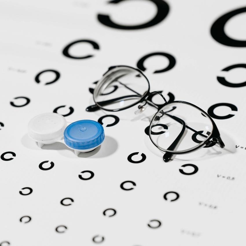 Contact lens case with a blue and white cap, next to a pair of eyeglasses on a vision test chart.