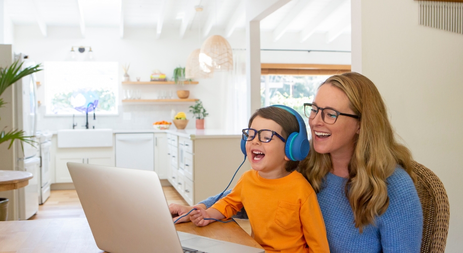 Child wearing headphones and an orange shirt smiling alongside an adult wearing glasses, looking at a laptop.