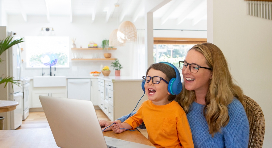 Child in orange shirt and blue headphones, sitting with a woman in glasses, smiling at a laptop.