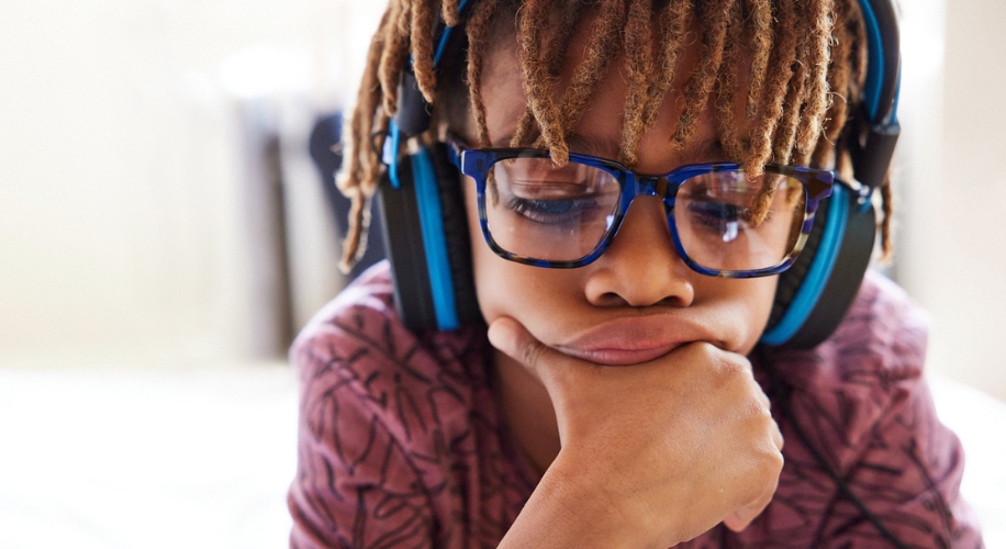 Child wearing oversized blue glasses and black headphones, looking thoughtful with chin on hand.