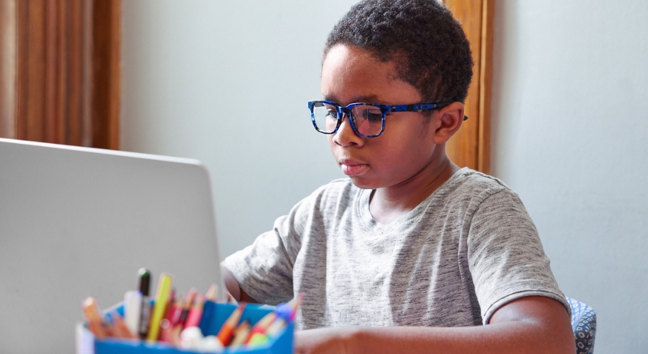 Child working on a laptop, wearing blue glasses and a gray shirt.