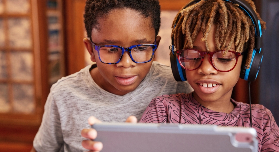 Two children looking at a tablet, one wearing blue glasses, the other with red glasses and headphones.