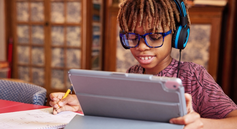 Young person using a tablet with a pencil and notebook, wearing blue headphones and glasses.