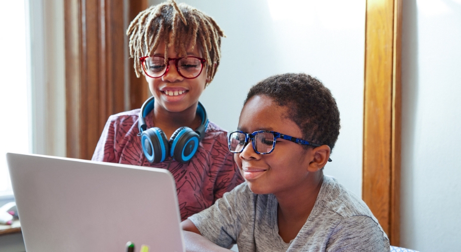 Two children wearing glasses using a laptop. One wearing blue headphones around the neck.