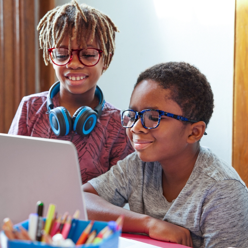 Two children wearing glasses, with one child also wearing blue headphones, looking at a laptop screen.