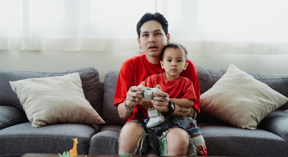 Man and young boy sitting on a couch, both wearing red shirts, holding a white game controller.