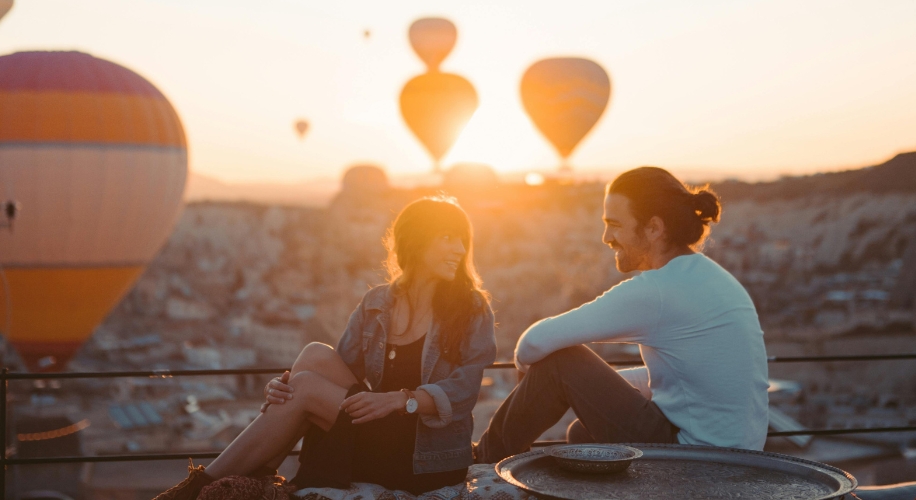 Two people sitting and smiling at each other with hot air balloons floating in the sunrise background.