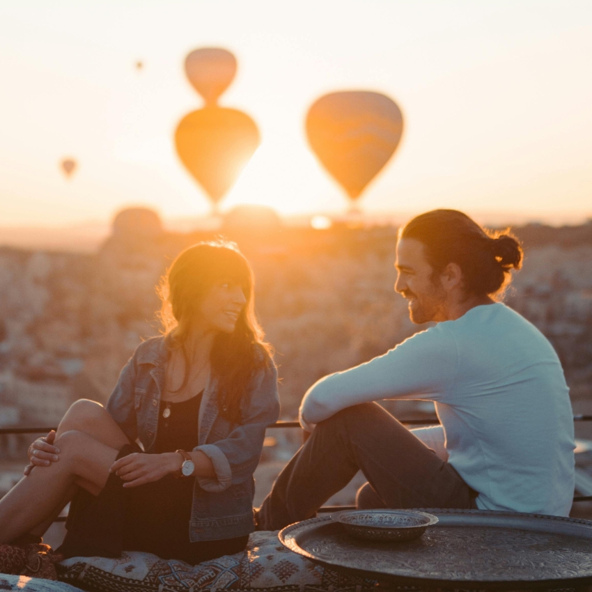 Couple sitting together at sunset with hot air balloons in the background.