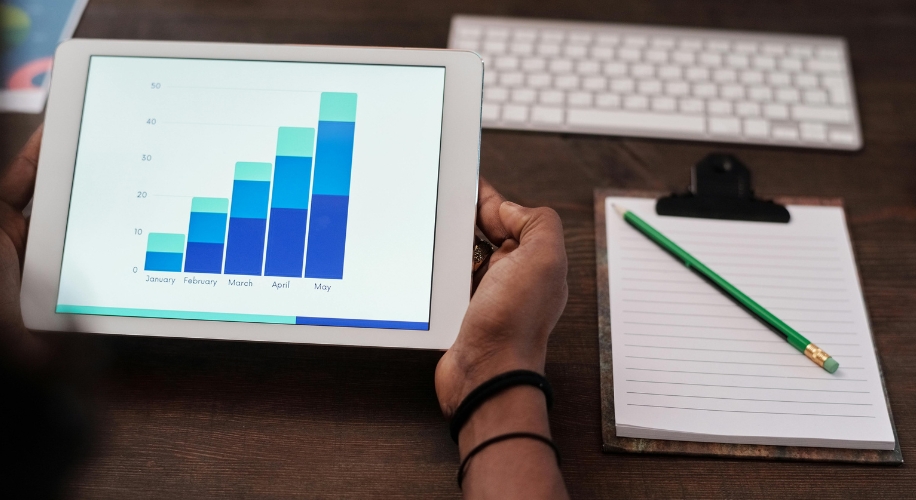 Tablet displaying a bar chart with data for January to May, held by a person next to a notepad and pencil.