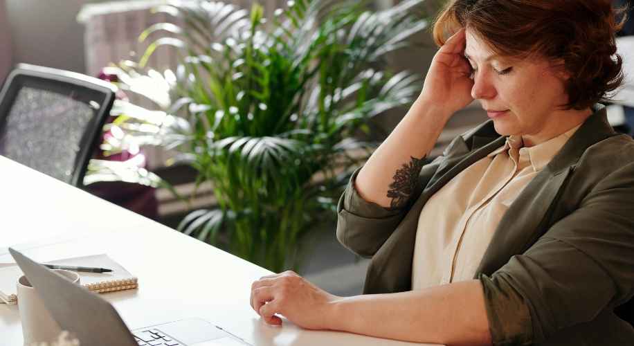 Person in a beige shirt and green jacket with one hand on the desk and the other against their temple.