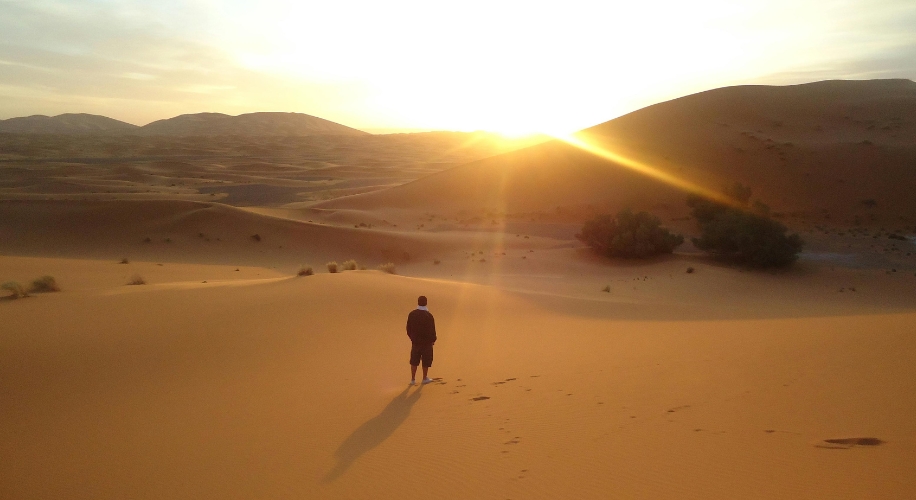 A person walking in a vast desert landscape with the sun setting behind distant dunes.