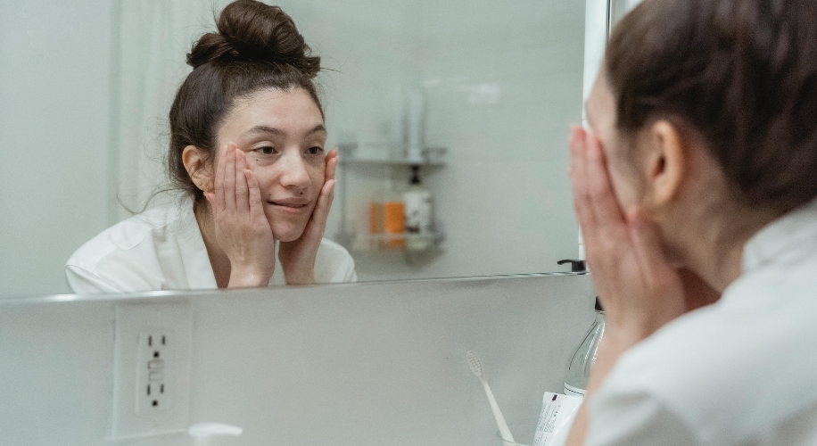 Woman with hair in a bun looking in the mirror, touching her face with both hands, wearing a white robe.