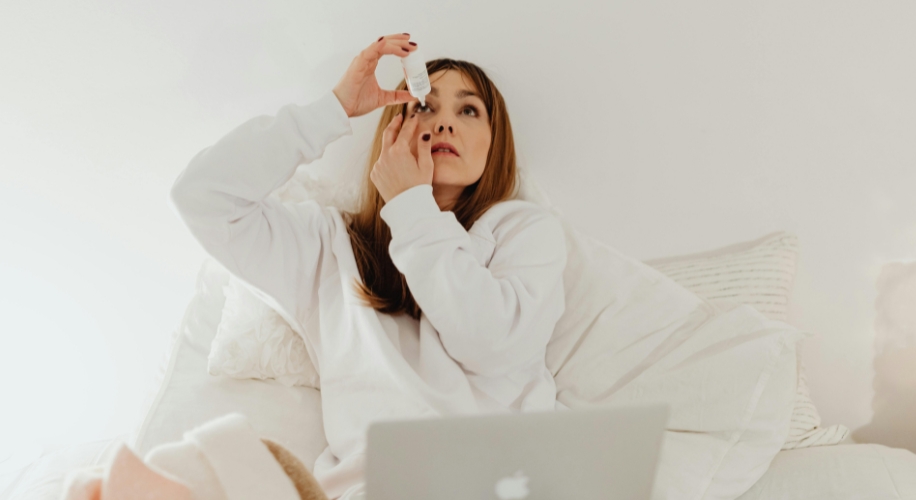 Woman applying eye drops while sitting on a bed with a laptop.