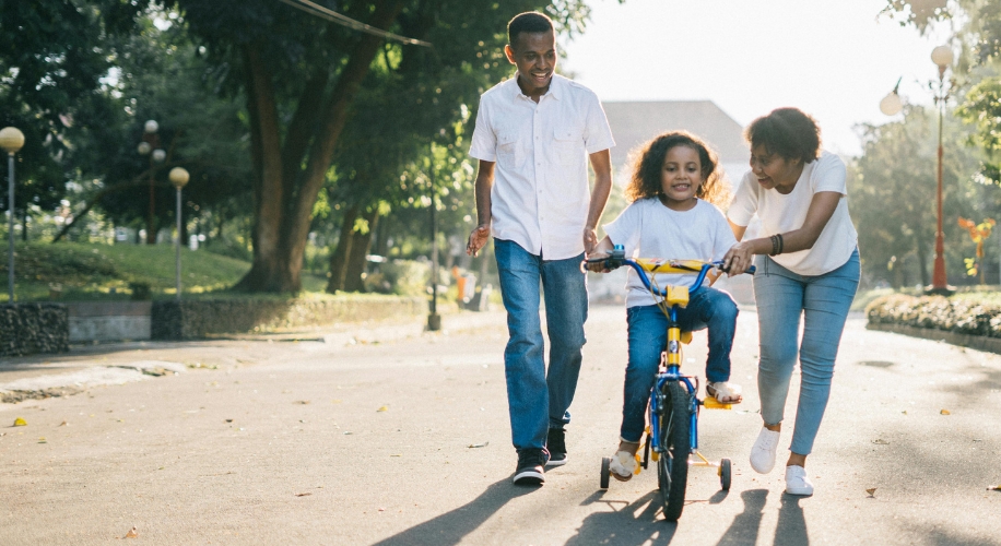 Child riding a bike with assistance from an adult man and woman on a sunny path surrounded by trees.