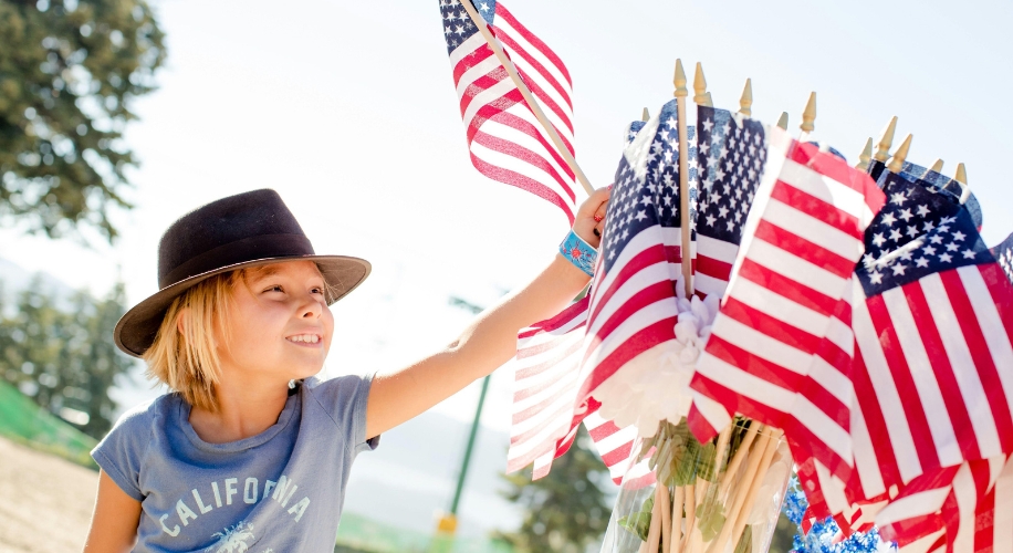 Child holding a small American flag, wearing a black hat and a blue &quot;California&quot; T-shirt.