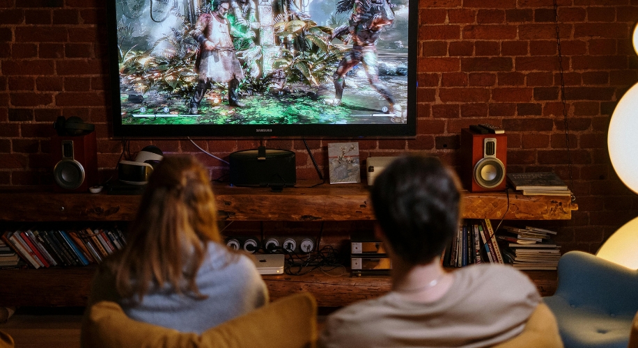 Two people sit on a couch playing a video game on a large wall-mounted TV.