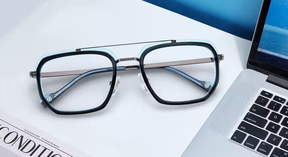 Square prescription glasses with black and blue frames, resting on a magazine near a laptop.