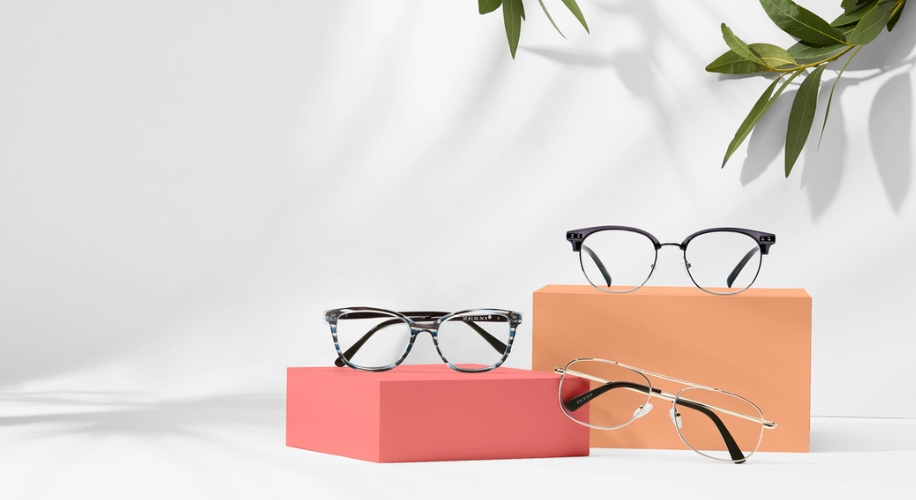 Three pairs of eyeglasses displayed on orange and pink blocks with leafy shadows.