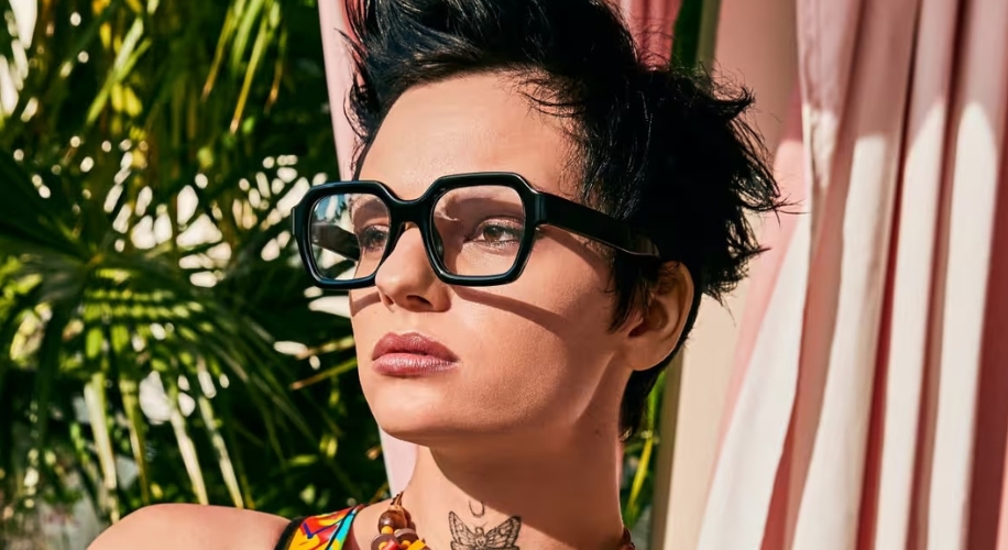 Person wearing large, square, black-rimmed glasses, colorful outfit, and necklace. Background has plants and curtains.