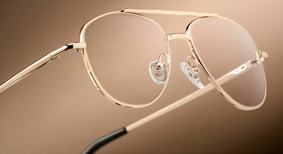 Gold-framed aviator-style glasses with clear nose pads, set against a beige background.