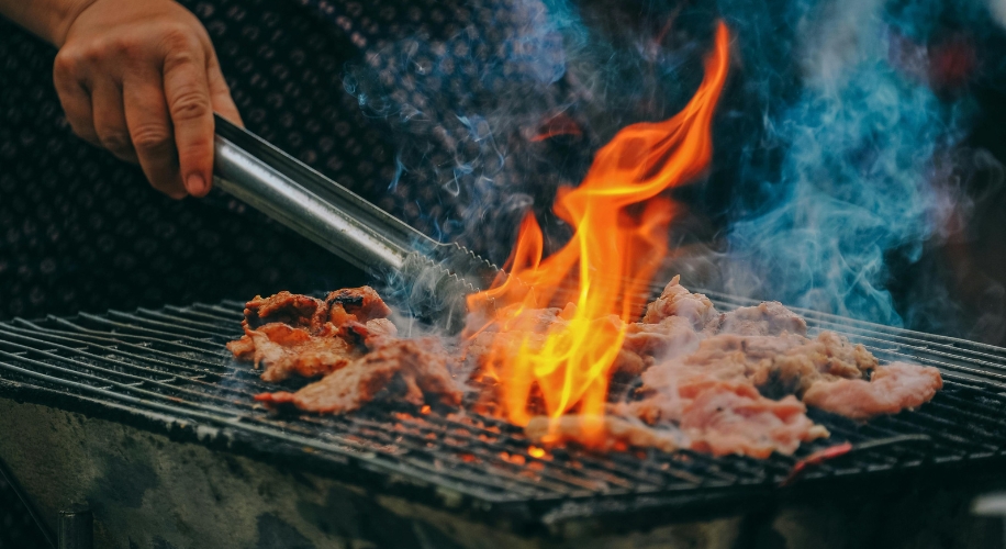 Grilling meat on a barbecue grill with flames and smoke, hand holding tongs turning the meat.