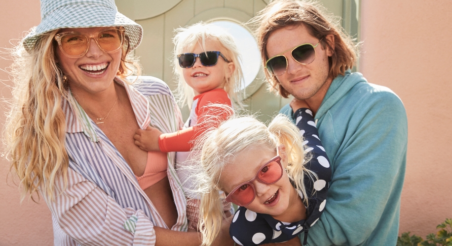 Family wearing different styles of sunglasses, smiles and embraces in front of a light green door.