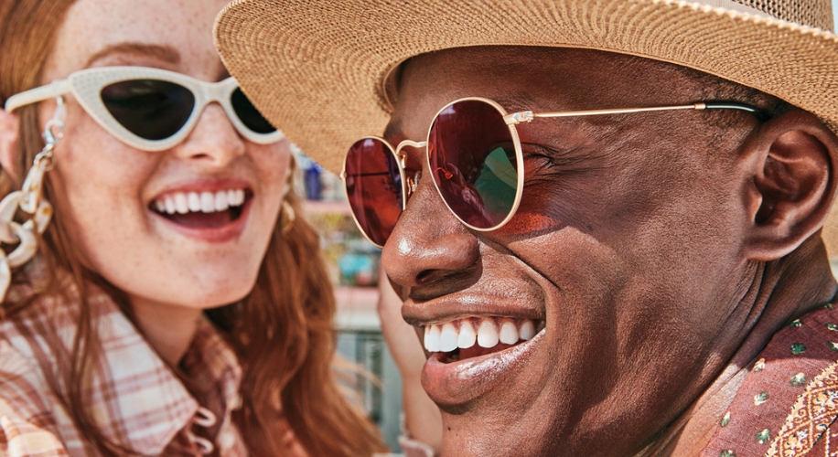 Two people smiling wearing stylish sunglasses; one with cat-eye frames, the other with round, tinted lenses.