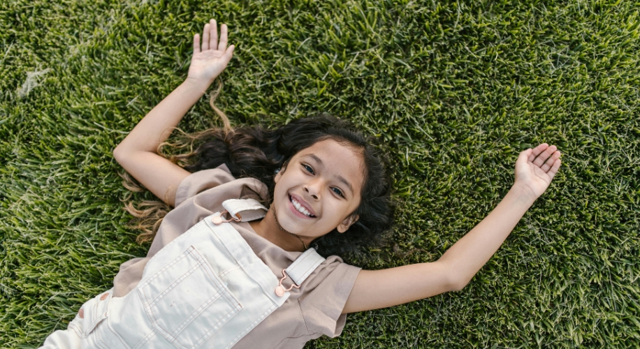 A smiling girl lying on green grass wearing a beige shirt and white overalls.