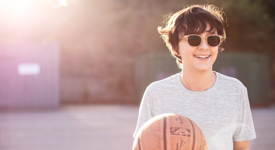 Teen wearing sunglasses, holding a basketball, and smiling in a sunlit outdoor scene.