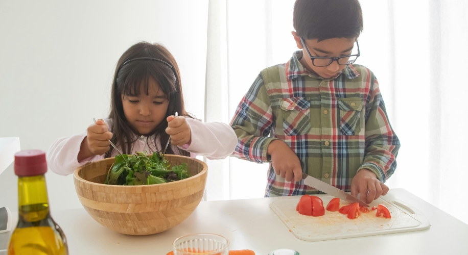 Two children preparing food; one mixing a salad, the other chopping tomatoes on a cutting board.