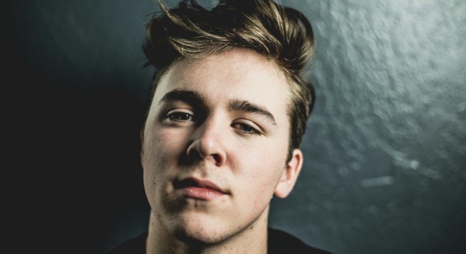 Young man with styled hair and neutral expression against a dark textured background.