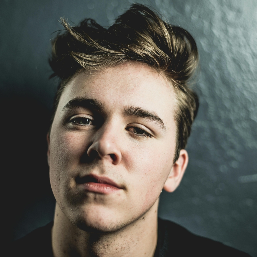 Close-up of a young man with light brown hair styled upwards, neutral expression, wearing a dark shirt.