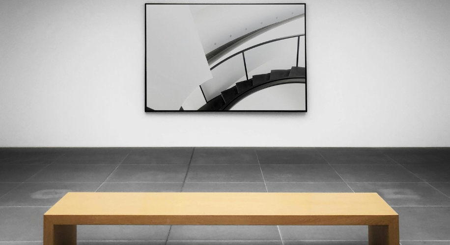 Framed abstract artwork of a spiral staircase, displayed on a white wall above a wooden bench.