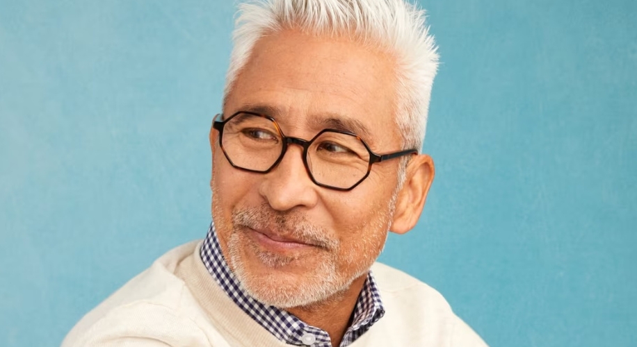 Man wearing black-framed glasses, checkered shirt, and beige sweater, smiling against a blue background.