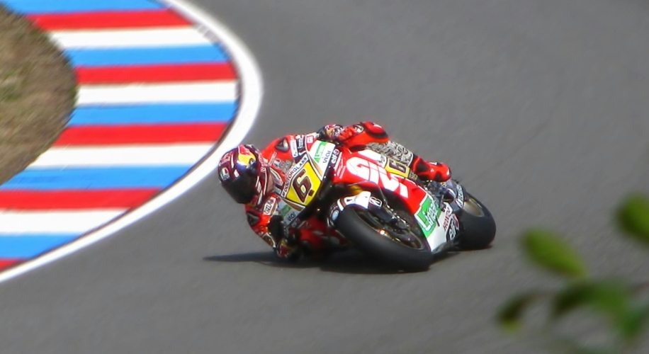 Motorcycle racer leaning into a turn, wearing a red and yellow suit, bike number 65 visible, with sponsor logos.