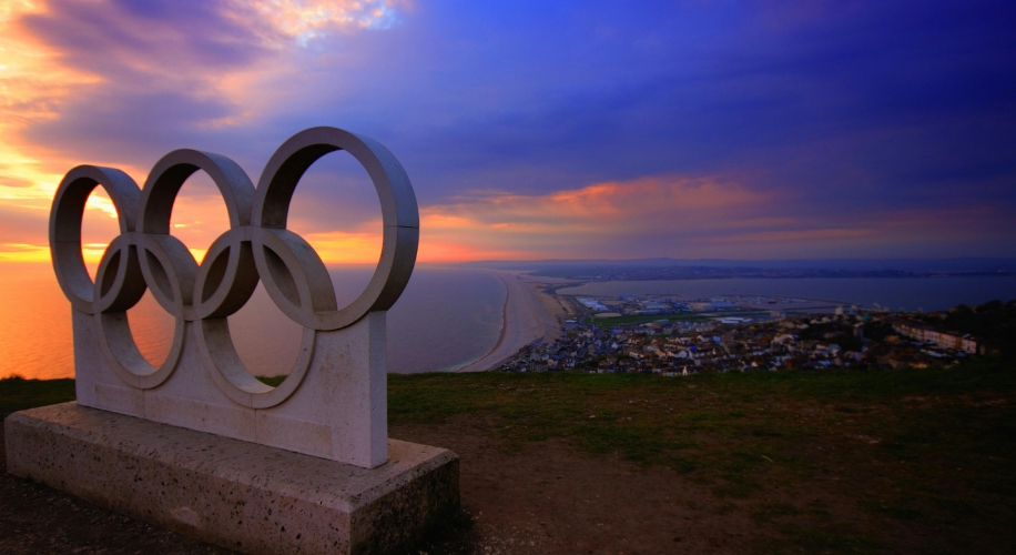 Olympic rings sculpture overlooking a coastal city at sunset.