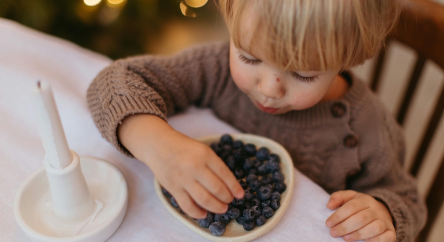 Child in a brown sweater eating blueberries from a small white dish.