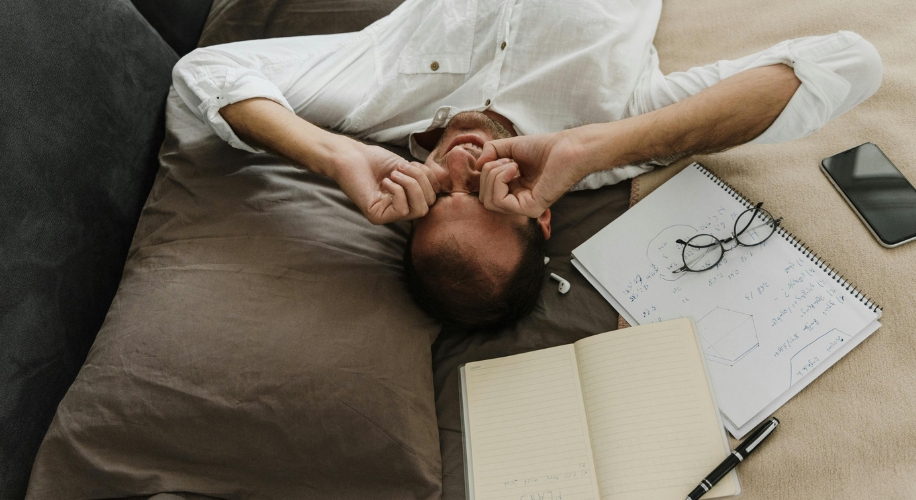 Man lying on a bed with notepad, spiral-bound book, phone, glasses, and pen.