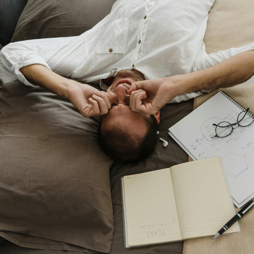 Man lying on a bed with hands over his face, next to a notebook labeled "PLANS" and a spiral notebook.