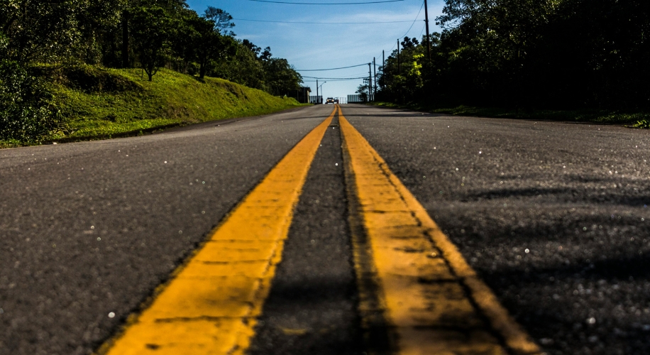 Road with two yellow lines extending into the distance on a sunny day. Trees and power lines in the background.