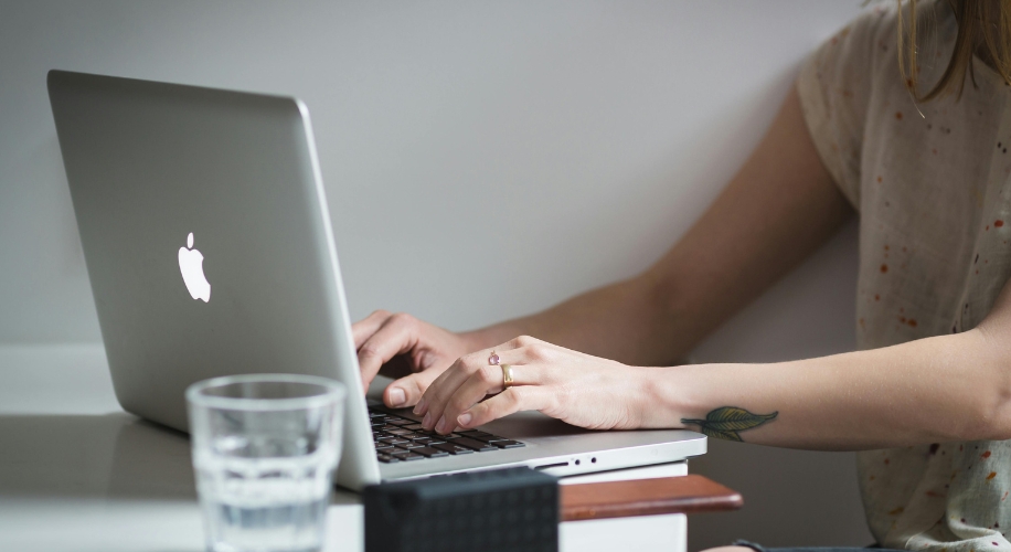 Person typing on a silver MacBook, with an Apple logo, placed on a white desk.