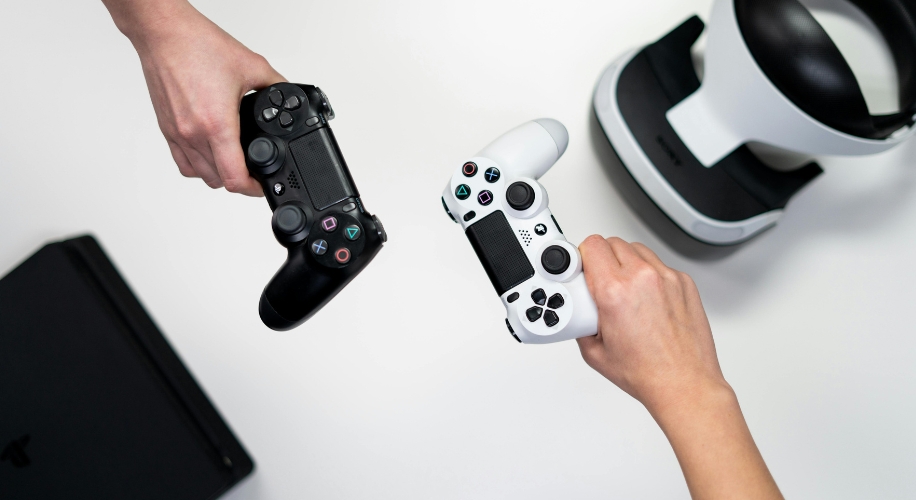 Two hands holding black and white PlayStation controllers, VR headset, and PS4 console in the background.