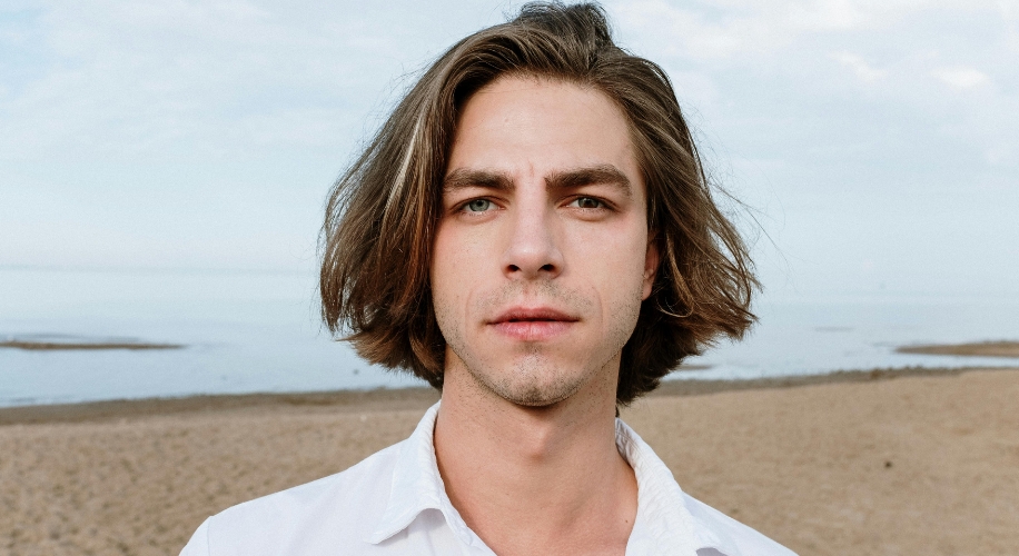 Young man with medium-length brown hair and a white shirt standing on a beach.