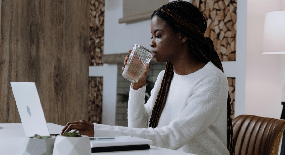 Woman in white sweater drinking water while using a laptop at a desk.